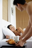 Woman in bed, man setting down breakfast tray on bedside table - Asia Images Group