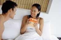Couple in bed, woman holding gift box, smiling - Asia Images Group