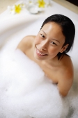 Woman sitting in bathtub, covered with soap suds, smiling up at camera - Asia Images Group