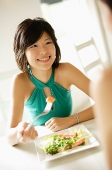 Young woman at table, eating salad, smiling at person in front of her - Asia Images Group