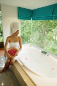 Woman in towel, sitting at edge of bath tub, holding bowl of flower petals - Asia Images Group
