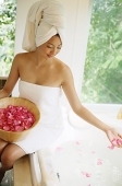 Woman in towel, sitting at edge of bath tub, throwing flower petals into water - Asia Images Group