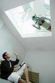 Man in living room, waving at woman in swimming pool through the skylight - Asia Images Group