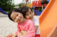 Two young girls sitting on slide, embracing, smiling at camera - Asia Images Group