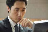 Businessman sitting at desk, looking at computer monitor, hand on chin - Asia Images Group