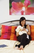 Girl sitting on bed, hugging stuffed toy, using mobile phone - Asia Images Group