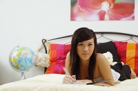 Girl lying on bed, holding pen, looking at camera - Asia Images Group