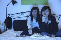 Two girls in bedroom, watching TV and eating popcorn - Asia Images Group