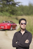 Man wearing sunglasses, arms crossed, red sports car in the background - Asia Images Group