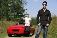 Man wearing sunglasses, facing camera, woman and red sports car in the background - Asia Images Group