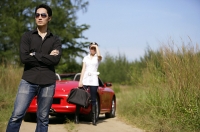 Man standing with arms crossed, woman and red sports car in the background - Asia Images Group