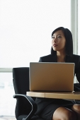 Businesswoman sitting at desk with laptop, looking away - Asia Images Group