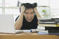 Young adult with laptop and books, hands in hair, grimacing - Asia Images Group