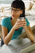 Young woman in cafe, holding cup, laptop on table next to her, looking away - Asia Images Group