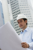 Man in hardhat, holding blueprints, building in the background - Asia Images Group