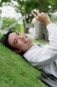 Businessman lying on grass in park, holding mobile phone, smiling at camera - Asia Images Group