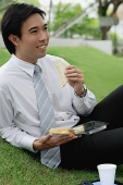 Businessman sitting on grass in park, having sandwich lunch - Asia Images Group