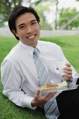 Businessman sitting in park, having take away lunch, looking at camera - Asia Images Group
