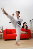 Couple at home, man practicing martial arts, woman sitting on sofa, filing her nails - Asia Images Group