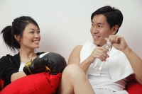Couple sitting side by side, woman with boxing gloves, man holding bottled water - Asia Images Group