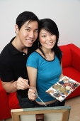 Couple at home, standing in front of easel with palette and paint brushes, smiling at camera - Asia Images Group