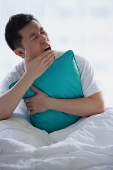 Man sitting in bed, embracing pillow, yawning - Asia Images Group