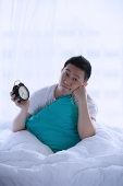 Man sitting in bed, holding alarm clock, hand on chin - Asia Images Group