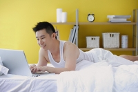 Man lying on bed in bedroom, using laptop - Asia Images Group