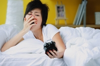 Woman in bed, looking at alarm clock, yawning - Asia Images Group