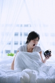 Woman in bed, looking at alarm clock, mouth open - Asia Images Group