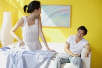 Couple at home, woman ironing turning to look at man sitting on sofa behind her - Asia Images Group