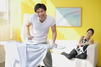 Couple at home, man ironing, woman sitting on sofa behind him - Asia Images Group