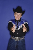 Senior man dressed in cowboy attire, pointing fingers at camera - Asia Images Group
