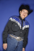 Senior man dressed in cowboy attire, standing against blue background - Asia Images Group