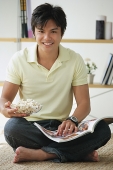Man sitting on floor, with magazine and bowl of popcorn - Asia Images Group