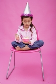 Girl with party hat, sitting on chair, holding bowl of cake - Asia Images Group
