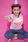 Girl with party hat, sitting on chair, holding bowl of cake towards camera - Asia Images Group