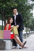Couple looking at camera, man standing, woman sitting, shopping bags around them - Asia Images Group