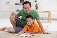Father and son in living room, smiling at camera, drawing materials around them - Asia Images Group