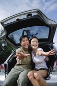 Couple sitting in boot of car, reaching towards camera - Asia Images Group