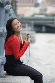 Woman sitting by river, listening to MP3 player, portrait - Asia Images Group
