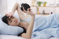 Young woman lying on bed, frowning at alarm clock - Asia Images Group