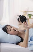 Young woman lying on bed, looking at alarm clock, yawning - Asia Images Group