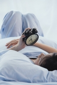Young woman lying on bed, looking at alarm clock - Asia Images Group