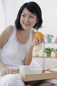 Young woman having breakfast in bed, holding orange, looking away - Asia Images Group
