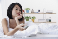 Young woman lying on bed, hand on chin, holding TV remote control - Asia Images Group