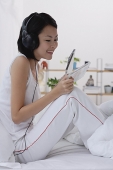 Young woman sitting on bed, listening to music, smiling - Asia Images Group