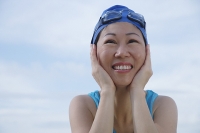 Mature woman wearing swim cap and goggles, hands on face, looking away - Asia Images Group