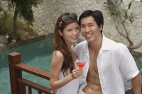 Couple standing side by side, smiling at camera, swimming pool behind them - Asia Images Group