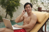 Man sitting by swimming pool, using laptop and mobile phone - Asia Images Group